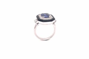One white gold ring stands upright, featuring a central blue gemstone encircled by small diamonds and a black embellishment. The ring's band has two small diamonds on either side of a mounted setting.