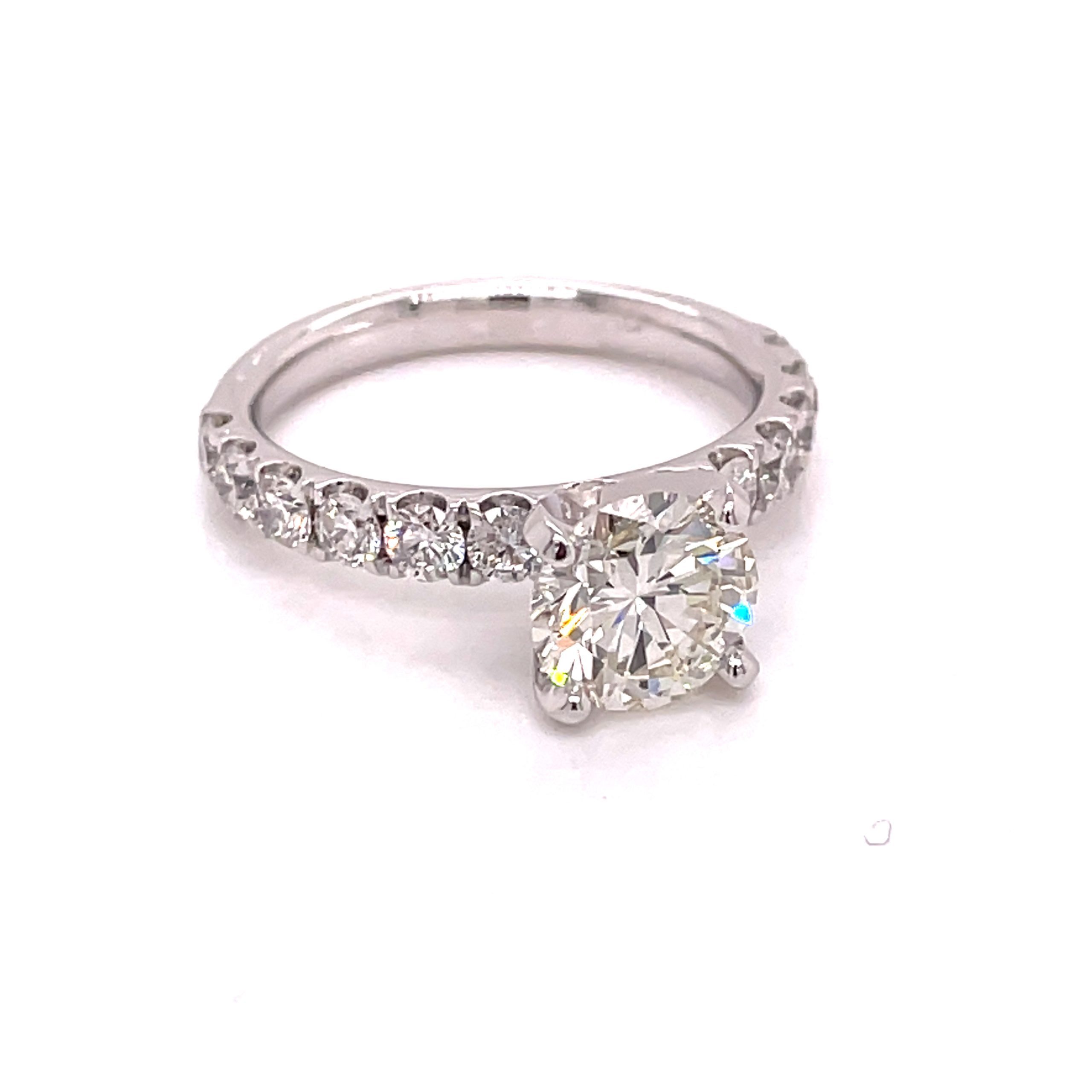 One white gold ring sitting on a white table. The ring feature a mounted round shaped diamond and the band of the ring is adorned with a round cut diamond.